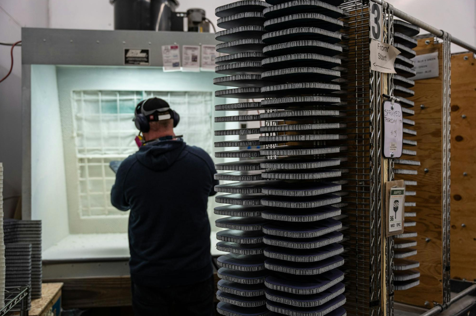 A man stands next to an 8-foot stack of unfinished pickleball paddles in a factory. 